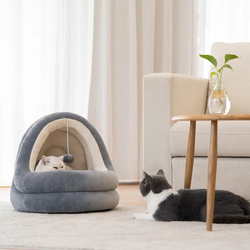 Un ambiente interior acogedor con dos gatos. A la izquierda, hay un gato acostado dentro de una cama para mascotas o una casa para gatos, que parece estar diseñada para brindar comodidad y calidez. El gato está mirando desde la cama de la mascota, posiblemente observando su entorno o al otro gato.