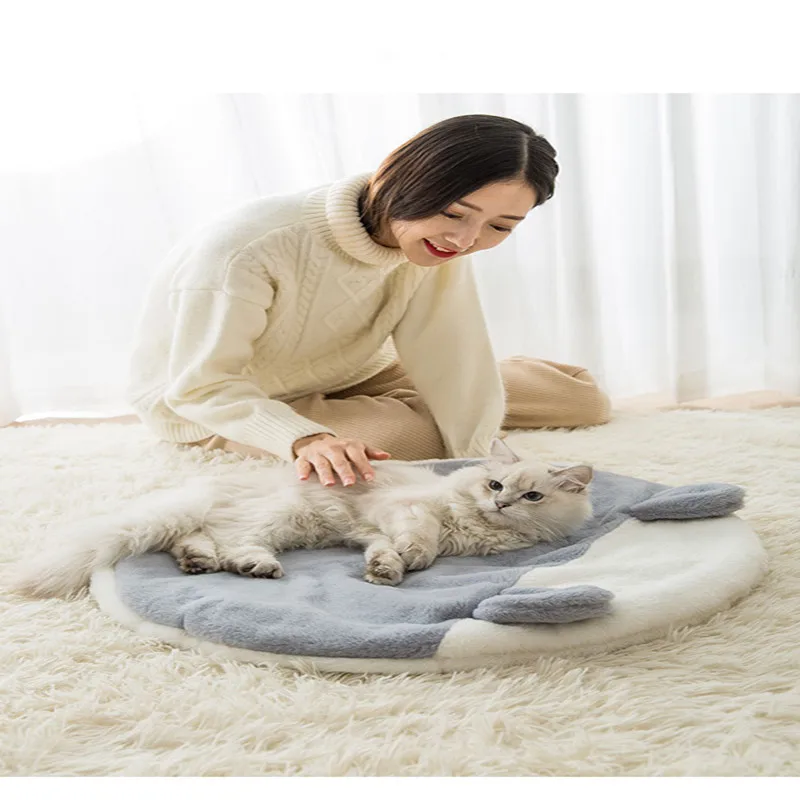 Una persona sentada en el suelo, sonriendo y acariciando a un gato blanco. El gato está acostado en una cama redonda y lujosa para mascotas con una combinación de colores azul y blanco. La persona viste un suéter blanco y parece estar disfrutando de un momento de relajación con el gato.