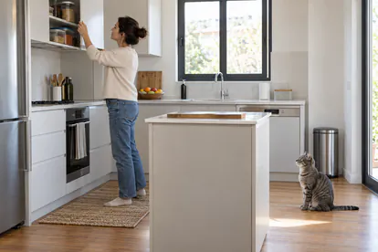 Persona que guarda alimentos en un armario alto mientras un gato espera en el suelo de la cocina
